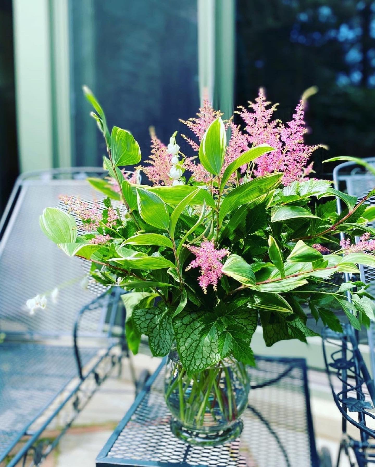 Bouquet of pink astilbe and green leaves in a clear vase on a metal table outdoors.